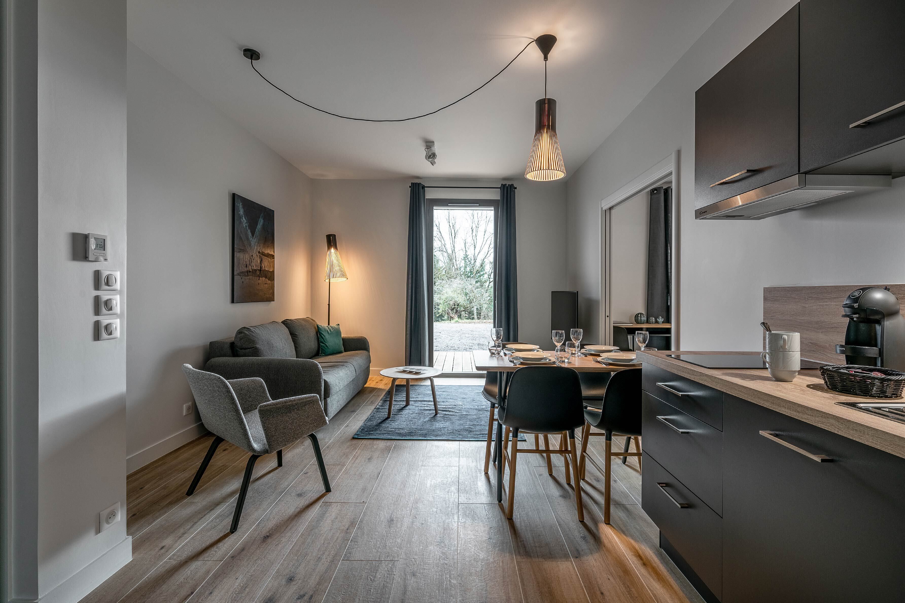 Eine moderne Küche mit dunklen Schränken, ein Essbereich mit Tisch und Stühlen, und ein graues Sofa mit Teppich.