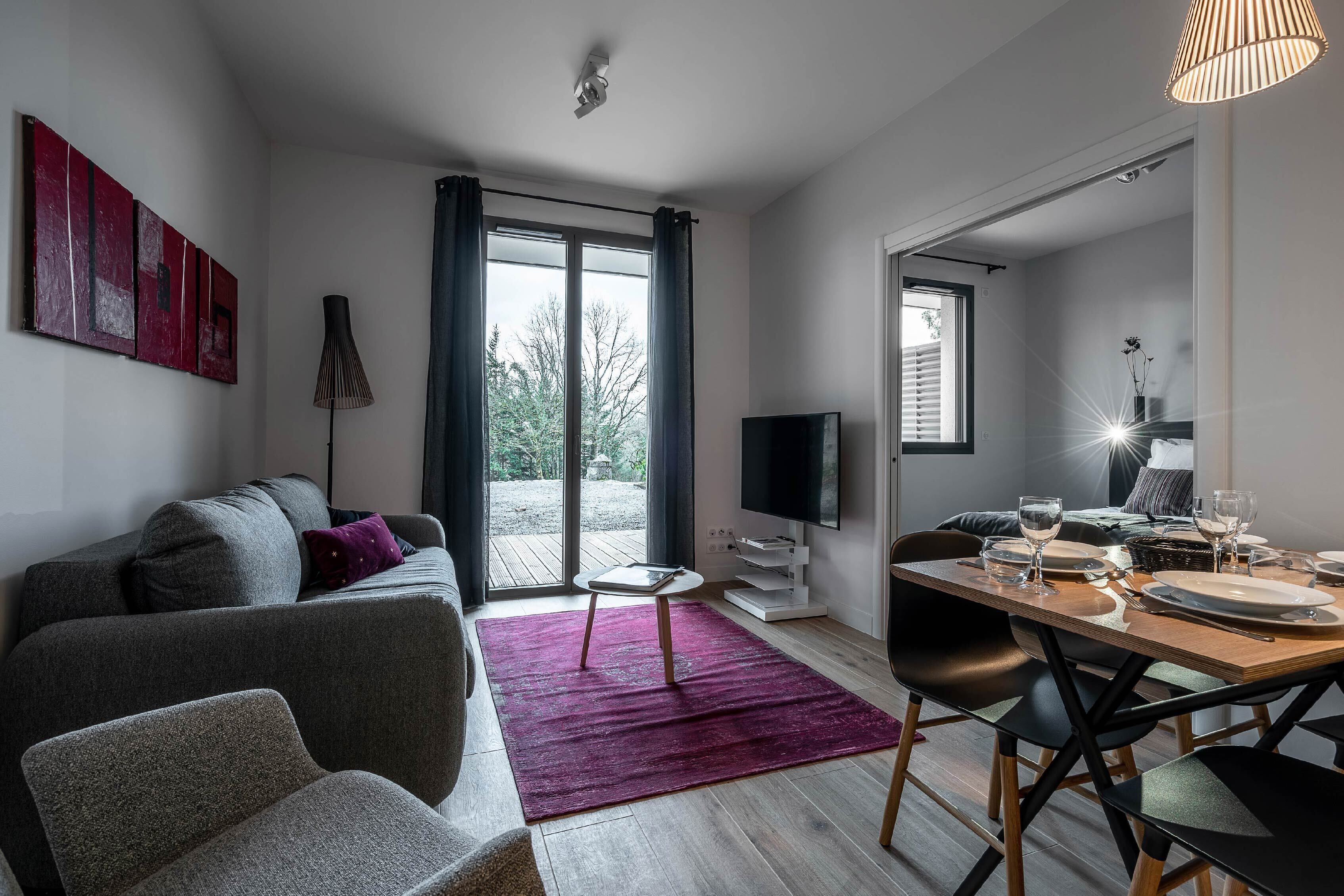 A modern living room with a grey sofa, a dining table set for four, and a large window with curtains.