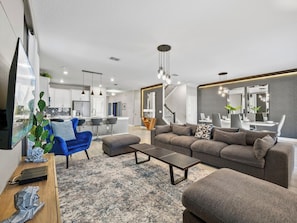 A modern living room with a sofa, ottoman, coffee table, and a blue armchair. There is a staircase leading to another level, and a kitchen area is visible in the background.