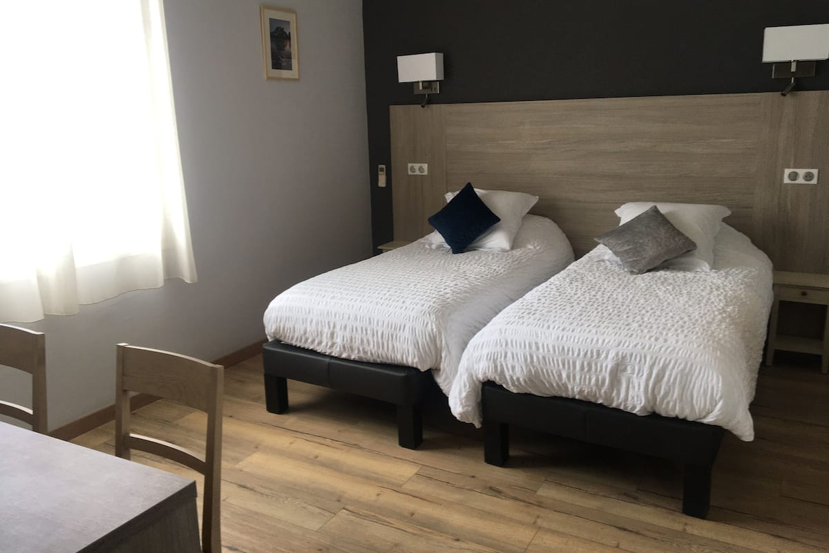 Quarto de hotel com duas camas, piso de madeira, escrivaninha e uma cadeira.