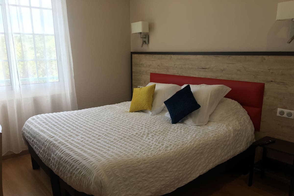 Cama bem arrumada com colcha branca texturizada e cabeceira vermelha. Três almofadas em amarelo, azul e branco. Uma mesa de cabeceira com controle remoto é visível.