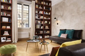 A living room with a sofa, a green ottoman, a coffee table, and two side tables. There are two large bookshelves filled with books and decorative items. A window with curtains is visible, and a lamp is on the wall.
