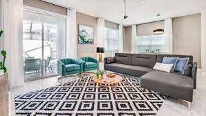 Una sala moderna con un sofá gris, sillones azul verdosos, un tapete estampado y un ventanal con persianas.