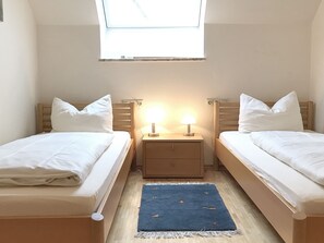 Quarto duplo com duas camas, uma mesinha de cabeceira, uma luminária, uma janela e um tapete azul.