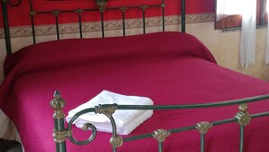 Uma cama com um cobre-leito vermelho e uma toalha branca, uma parede vermelha com uma pintura e uma janela com cortinas.
