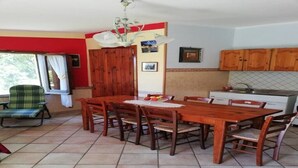 Uma área de refeições com uma mesa e cadeiras de madeira, uma poltrona verde e uma cozinha ao fundo.