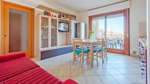 Ein modernes Wohnzimmer mit einem roten Sofa, einem Essbereich mit Tisch und Stühlen, einer Küche mit Kühlschrank sowie eine Schiebetür zu einem Balkon.