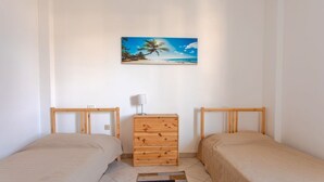 Ein Schlafzimmer mit zwei Einzelbetten, einer Holzkommode, einem Nachttisch mit Lampe und einem Wandbild mit Strandmotiv.