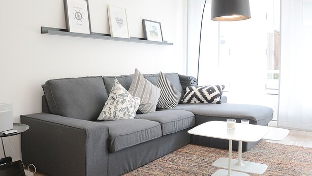 A modern living room with a grey sectional sofa, a white coffee table, and a black floor lamp. There are framed pictures on a grey shelf above the sofa. - Wohnung Gartenstraße 23 (Norderney)