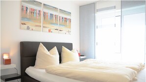 A neatly made bed with white linens and pillows, a bedside table with a lamp, and artwork on the wall. - Wohnung Gartenstraße 23 (Norderney)