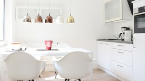 A modern kitchen with white cabinetry, a white dining table, and four pendant lights. - Wohnung Gartenstraße 23 (Norderney)