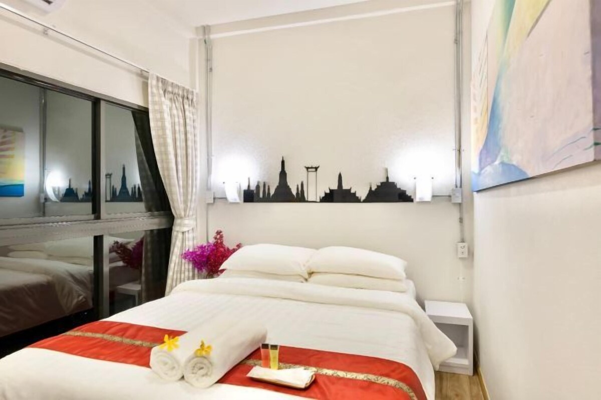 Een hotelkamer met een opgemaakt bed, een stadslandschap schilderij aan de muur, en een raam met gordijnen.