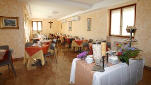 Un comedor con varias mesas dispuestas para una comida, con una estación de buffet que ofrece diversos platos y un centro de mesa floral.