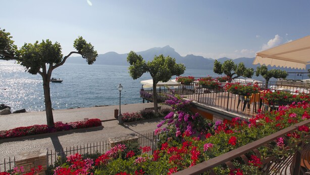 Un paseo marítimo con vistas al mar y a las montañas, que cuenta con una cafetería con terraza y macizos de flores.