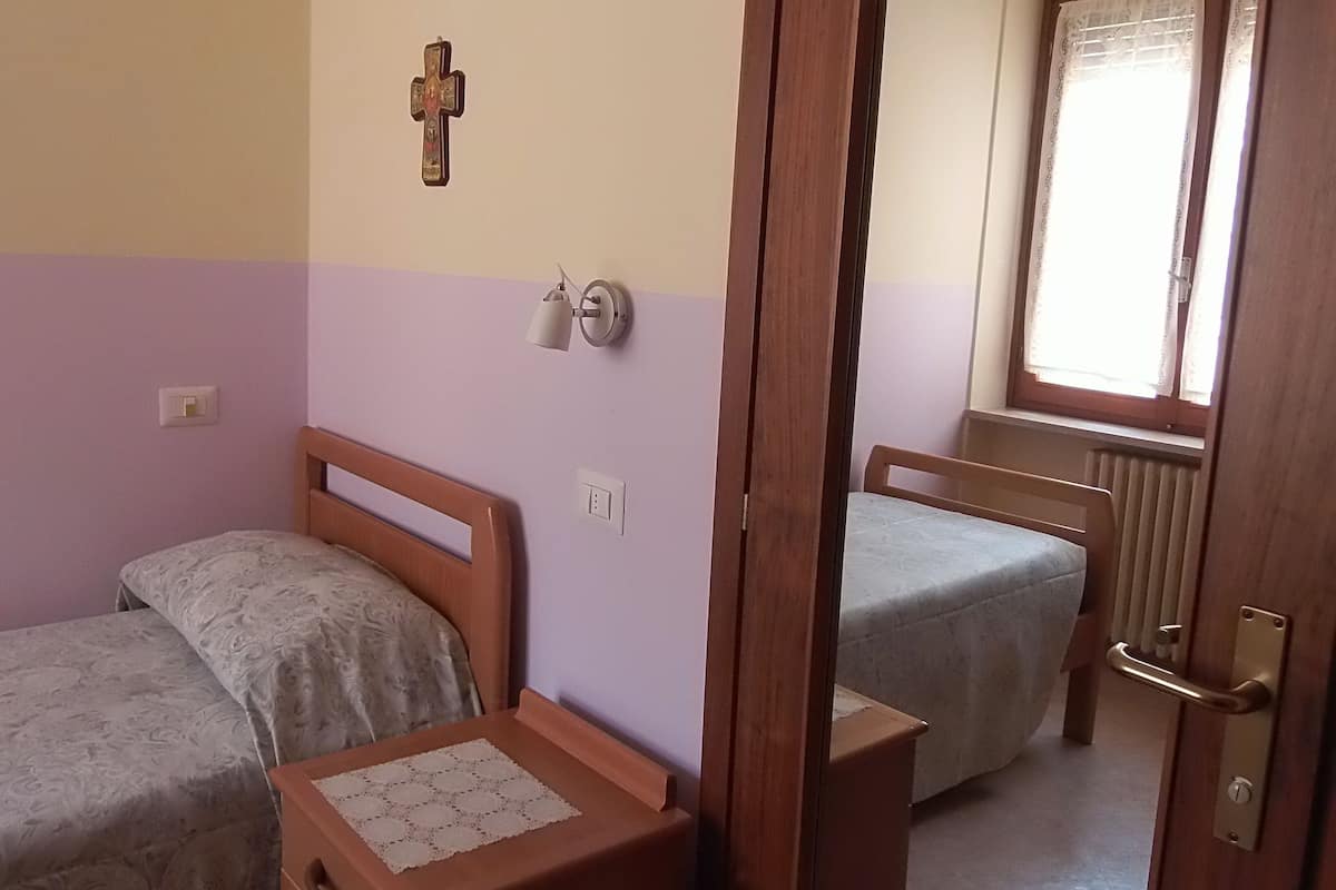 Een kamer met twee bedden, een houten deur, een raam met gordijnen en een aan de muur bevestigd kruisbeeld.