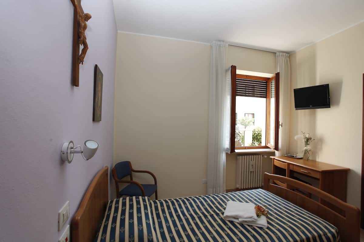 Een slaapkamer met een bed, een stoel, een bureau, een televisie en een raam met jaloezieën.