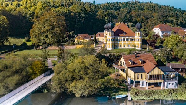 Ein großes, gelbes Gebäude mit mehreren Türmen, umgeben von üppiger Vegetation und einem Fluss.