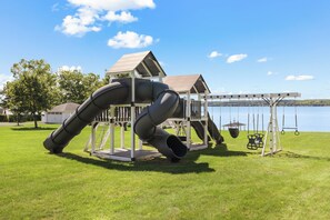 Un parque infantil con toboganes y columpios, con un lago y árboles de fondo.