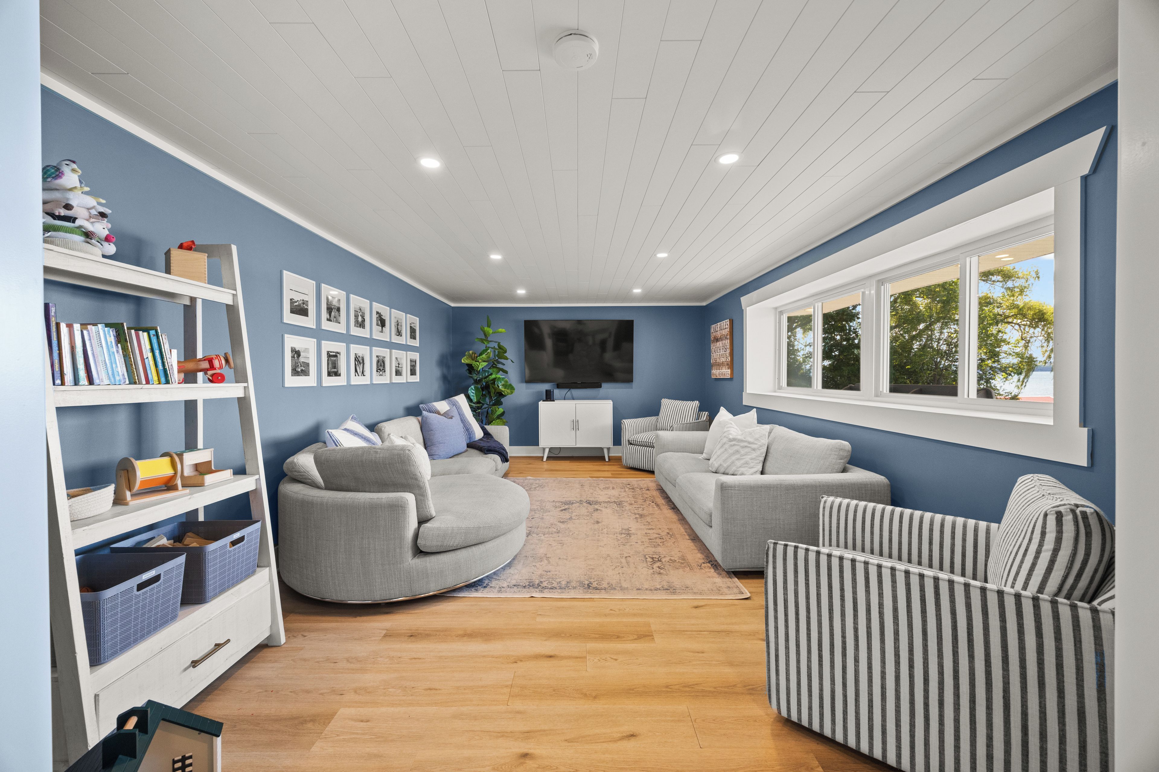 Ein Wohnzimmer mit blauer Wand, einem Flachbildfernseher, grauen Sofas, einem weißen Schrank und einem Bücherregal mit Büchern und Spielzeug.