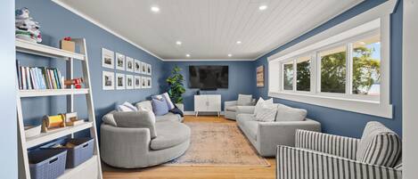 Ein Wohnzimmer mit blauer Wand, einem Flachbildfernseher, grauen Sofas, einem weißen Schrank und einem Bücherregal mit Büchern und Spielzeug.