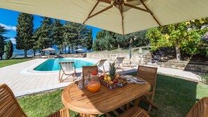 A poolside area with a table set for dining, surrounded by lounge chairs, and an umbrella providing shade. - Borgo Colle Ridente 13, Emma Villas (Camerino)