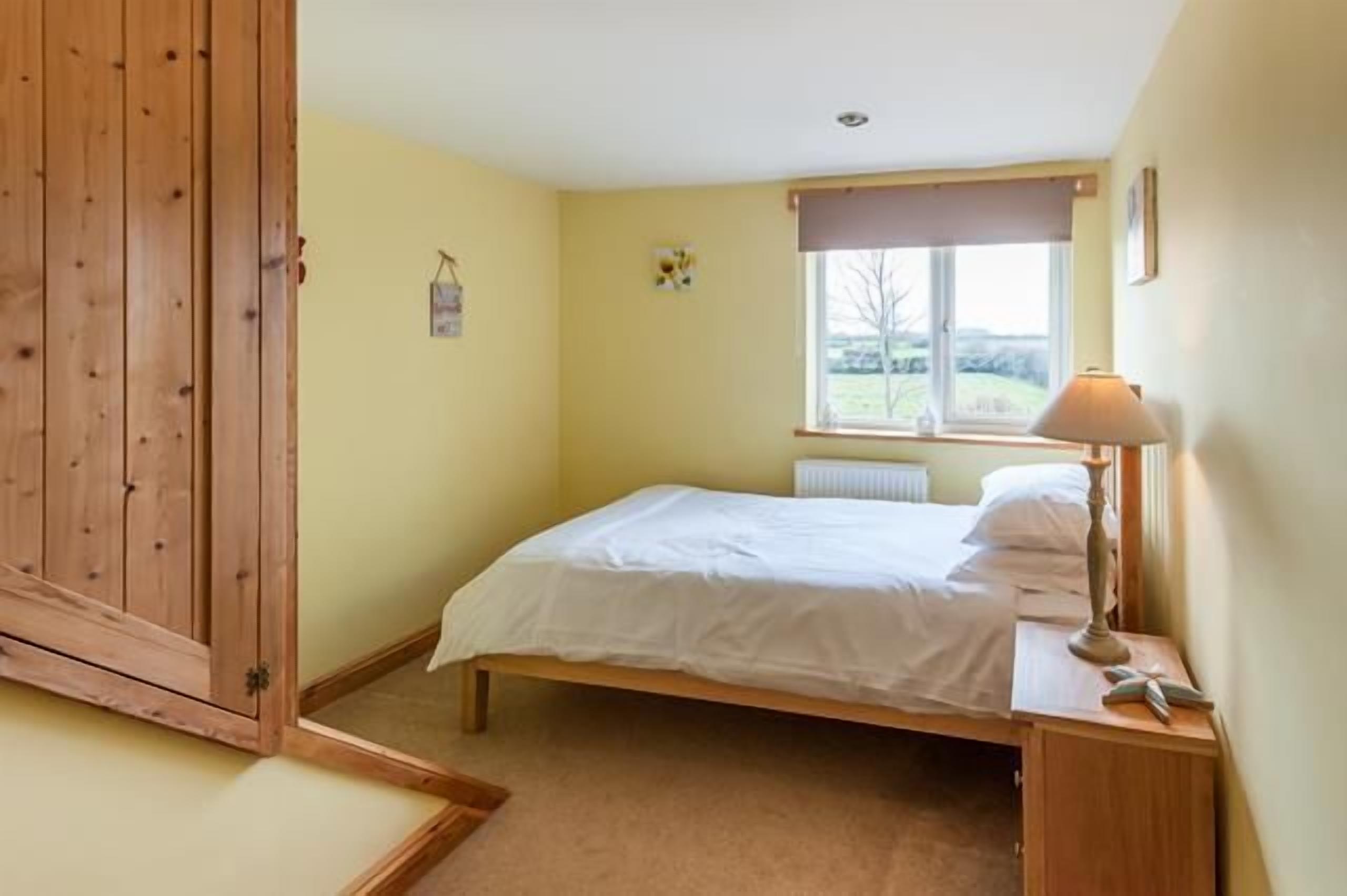 A bedroom with a single bed, a wooden wardrobe, a bedside table with a lamp, and a window with a view of the outdoors.