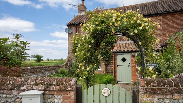 Une maison en briques avec une porte verte, un mur de pierres et un portail sur lequel est apposée une plaque.