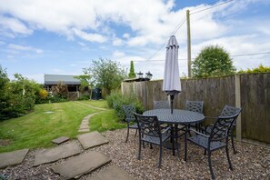 A patio with a round table and chairs, surrounded by a wooden fence and a garden path. - Corner Cottage | East Ruston Cottages. Happisburgh, Norfolk (Norwich)
