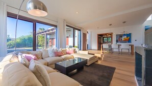 Amplia sala de estar con un sofá grande, una mesa de centro y una chimenea. Hay un comedor con sillas y una mesa, y ventanas amplias que ofrecen vistas al exterior.