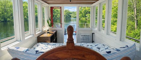 Cabina de barco con timón de madera, ventanas amplias, una mesita con una planta y vista a un lago.