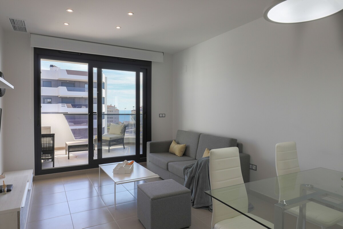 A modern living room with a glass table, grey sofa, and a view of the city through large sliding doors.