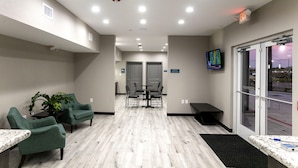A reception area with a reception desk, two green armchairs, a small table with a plant, a TV mounted on the wall, and a view of the outside through a glass door.
