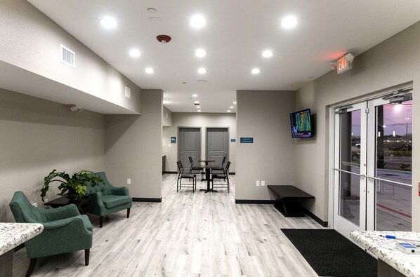 A reception area with a reception desk, two green armchairs, a small table with a plant, a TV mounted on the wall, and a view of the outside through a glass door.