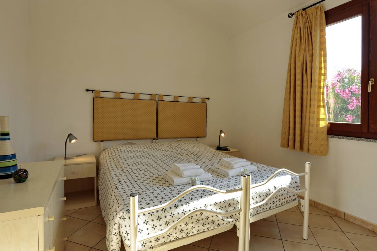 A neatly made bed with a patterned bedspread, a bedside table with a lamp, and a window with curtains.
