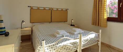 Una cama bien hecha con una colcha estampada, una mesita de noche con una lámpara y una ventana con cortinas.