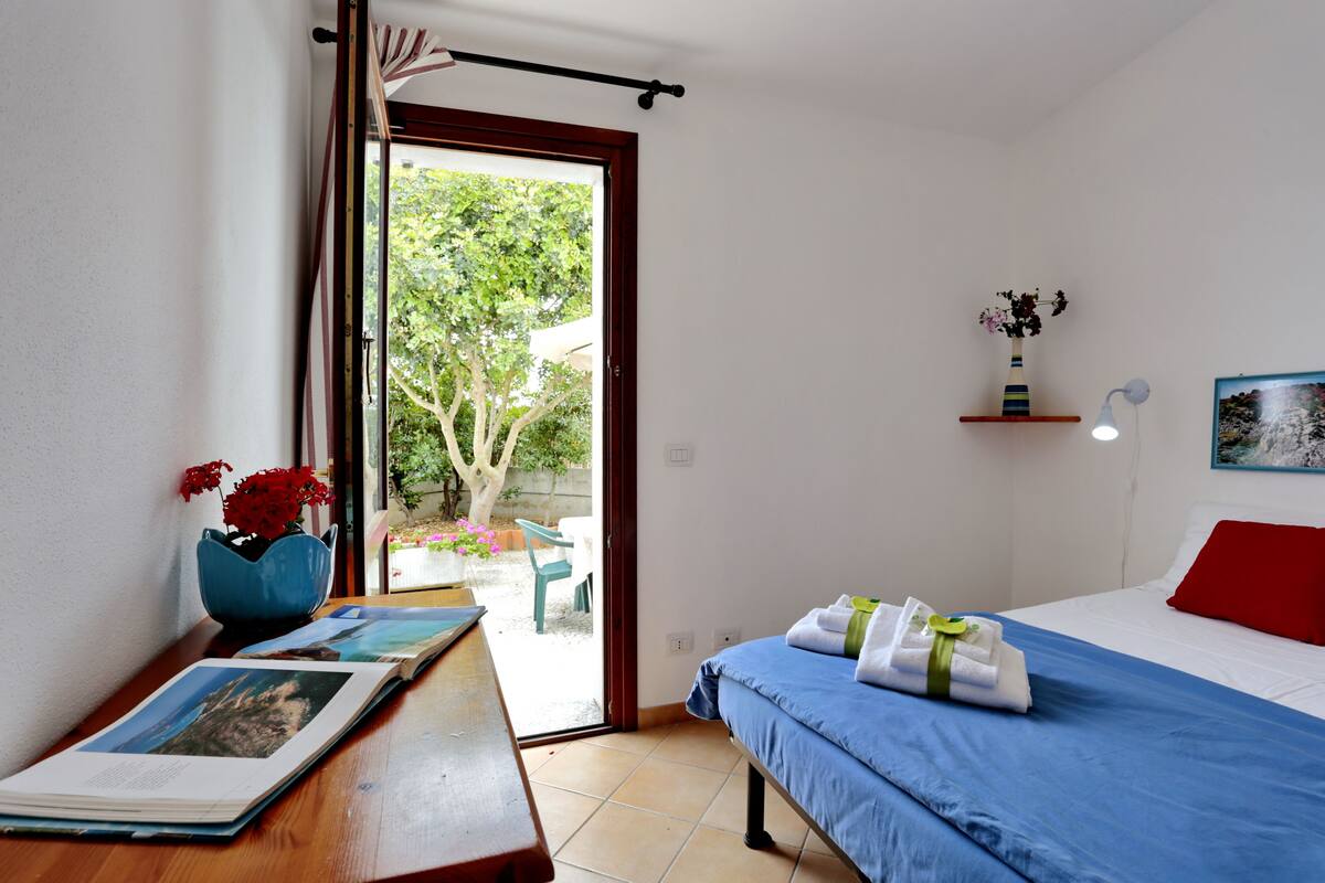 A hotel room with a bed, a desk with a book and a vase, a window with a view of trees, and a red pillow on the bed.