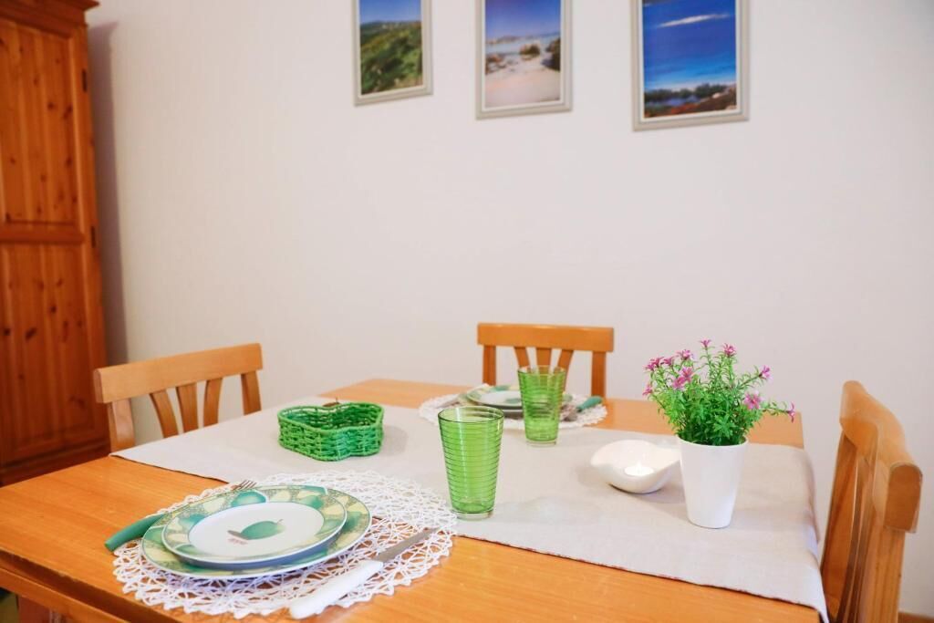 A dining table set with green glassware, plates, and a white napkin. There are two chairs, a wooden cabinet, and a potted plant with pink flowers.