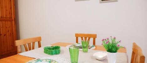 A dining table set with green glassware, plates, and a white napkin. There are two chairs, a wooden cabinet, and a potted plant with pink flowers.
