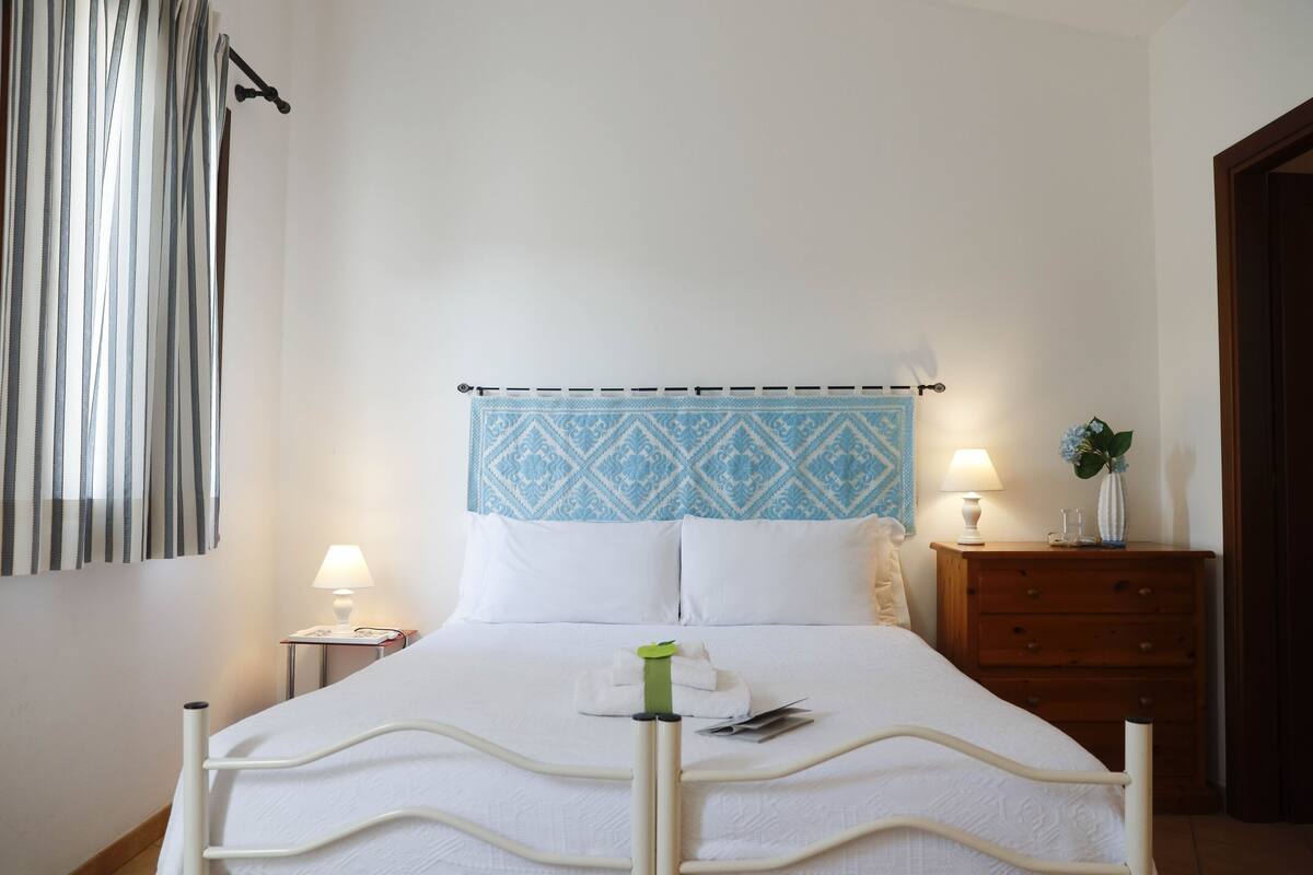 A neatly made bed with a patterned headboard, a bedside table with a lamp, and a wooden nightstand with a vase.