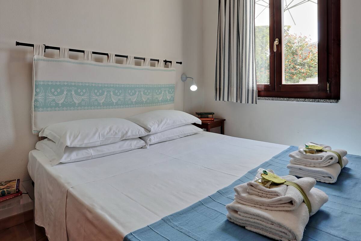 A neatly made bed with white linens, a blue bedspread, and two towels with green tags on a blue mat.