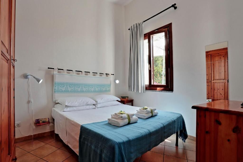 A bedroom with a bed, a desk, a window with curtains, a wall-mounted shelf, and a wooden door.
