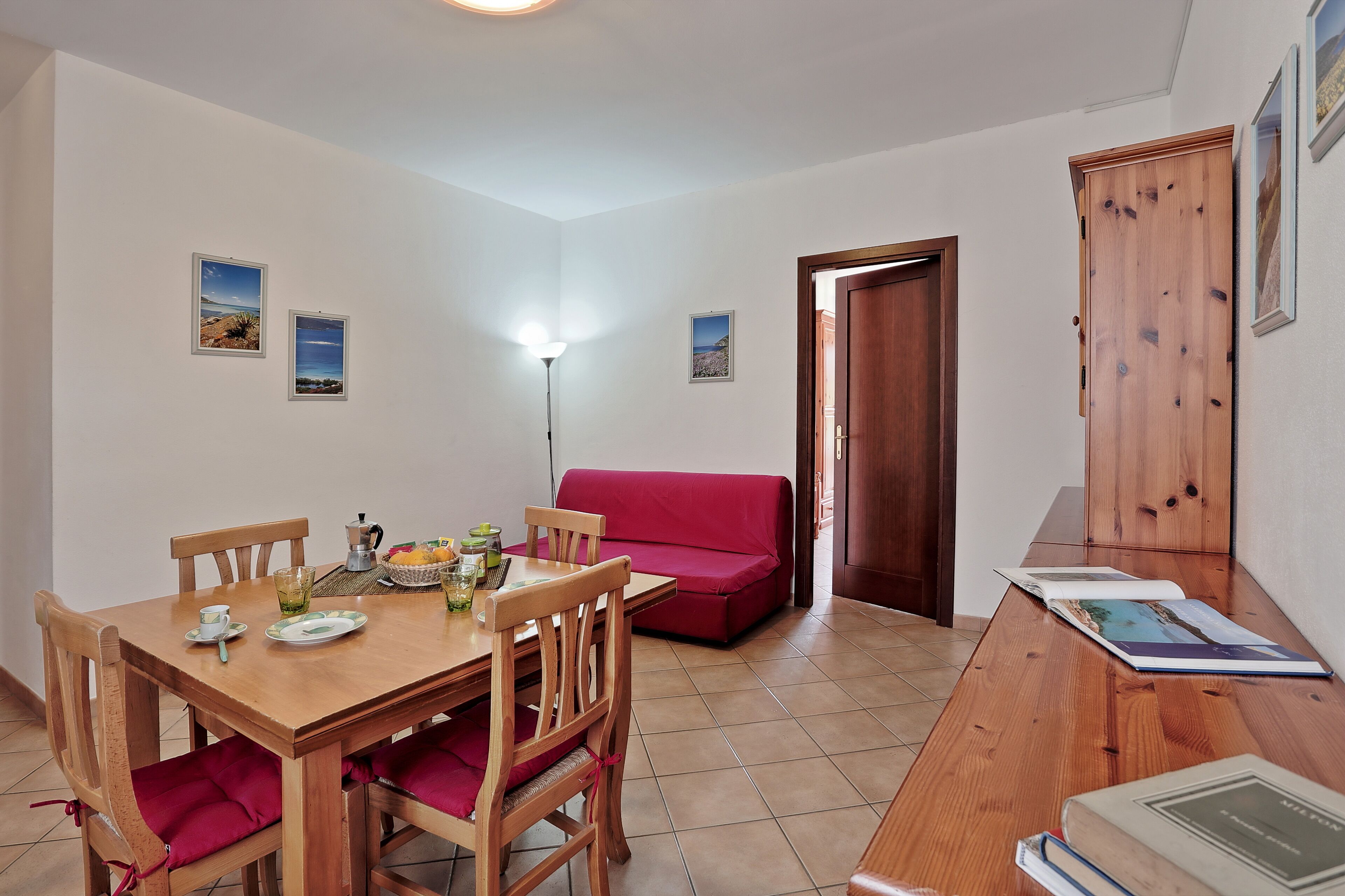 A dining area with a wooden table, red cushioned chairs, and a red sofa. There are framed pictures on the walls and a wooden cabinet.