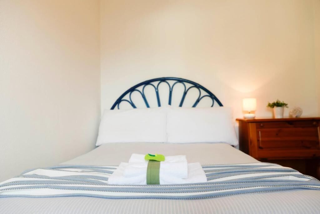 Una cama bien hecha con una colcha a rayas y almohadas, una mesita de noche de madera con una lámpara y una planta pequeña, y un cabecero de metal.