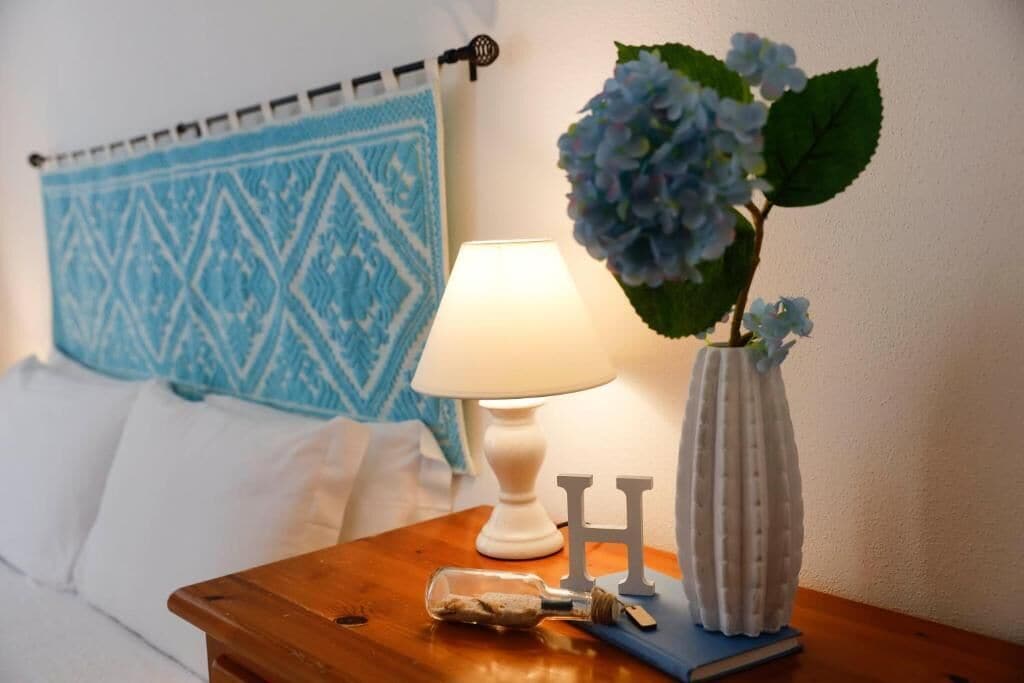 A neatly made bed with a blue patterned headboard, a bedside lamp, a vase with blue flowers, and a small bottle with a tag.