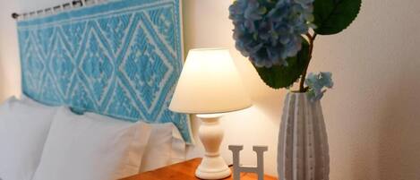 A neatly made bed with a blue patterned headboard, a bedside lamp, a vase with blue flowers, and a small bottle with a tag.