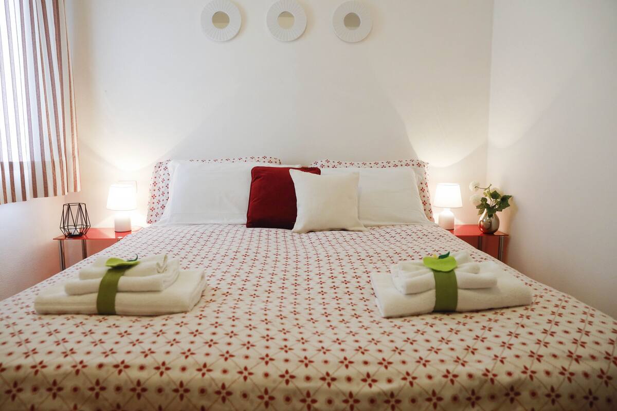 A neatly made bed with a patterned bedspread, two pillows, and two folded towels with a green ribbon.