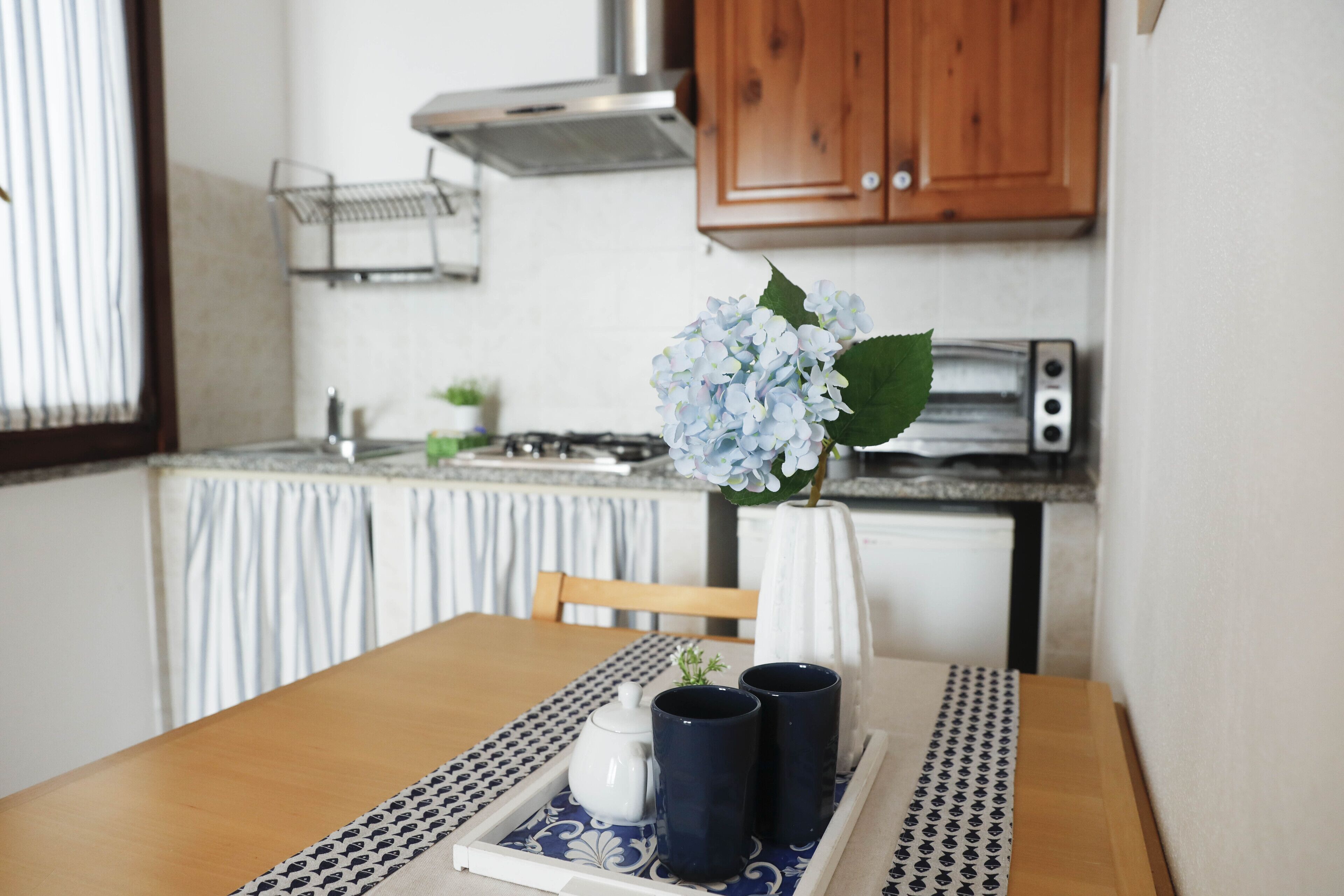 A dining table with a tray, two dark mugs, a white teapot, and a vase with blue flowers.