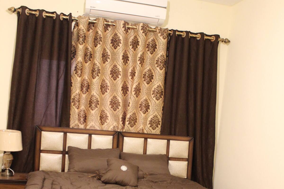 Une chambre à coucher avec une tête de lit en bois, une couvre-lit marron, un rideau à motifs et une lampe de chevet.