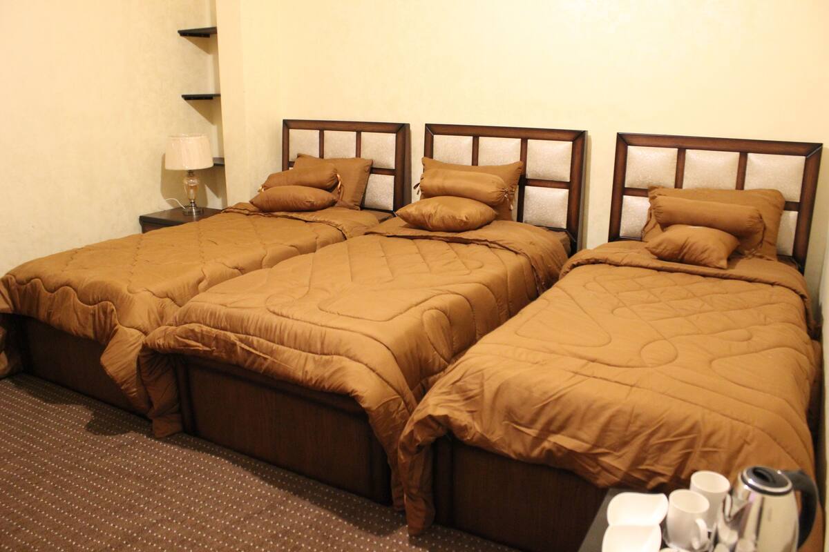 Une chambre d’hôtel avec trois lits simples, chacun doté d’une tête de lit en bois et de draps marron. On y trouve une petite table avec une lampe, ainsi qu’un plateau avec des tasses et une bouilloire.