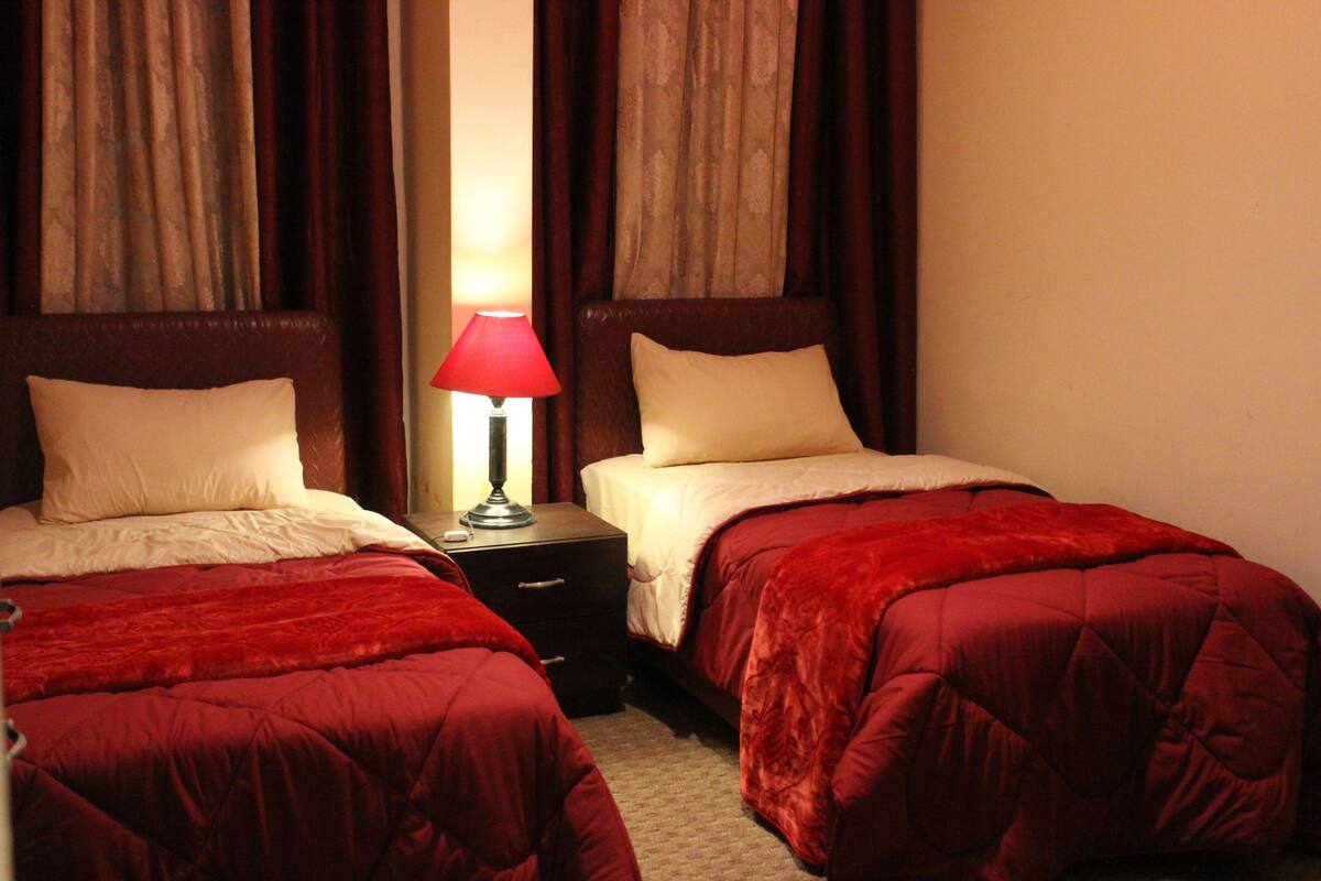Une chambre d’hôtel avec deux lits, chacun recouvert d’une couverture rouge et agrémenté d’oreillers blancs. Une table de chevet avec une lampe se trouve entre les deux lits.
